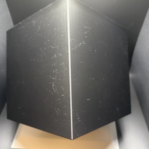 May include: A solid black cube with sharp, defined edges and a smooth, matte surface texture. The object is positioned at an angle, highlighting two visible faces that meet at a vertical seam, creating a geometric and symmetrical appearance.
