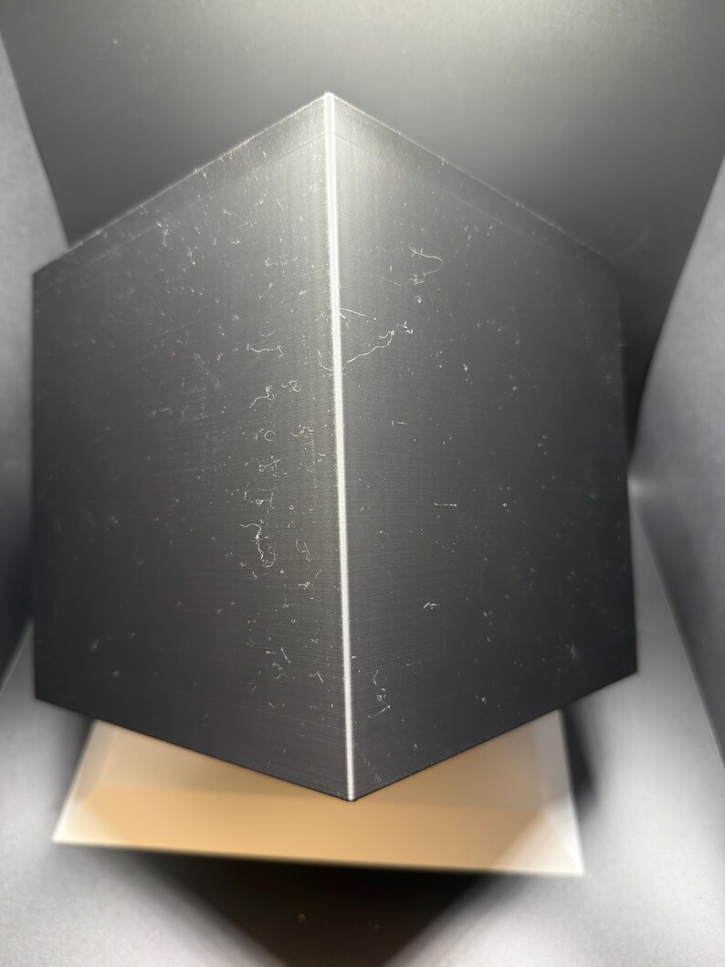 May include: A solid black cube with sharp, defined edges and a smooth, matte surface texture. The object is positioned at an angle, highlighting two visible faces that meet at a vertical seam, creating a geometric and symmetrical appearance.