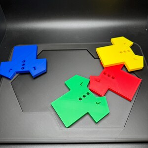 Puede incluir: Cuatro piezas de rompecabezas geom&eacute;tricas de colores azul, amarillo, rojo y verde est&aacute;n dispuestas sobre una superficie gris oscuro. Cada pieza tiene una forma &uacute;nica con tres agujeros circulares y una peque&ntilde;a muesca, lo que sugiere un rompecabezas o juego.