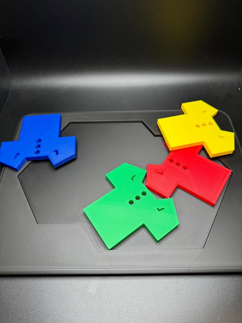 Puede incluir: Cuatro piezas de rompecabezas geom&eacute;tricas de colores azul, amarillo, rojo y verde est&aacute;n dispuestas sobre una superficie gris oscuro. Cada pieza tiene una forma &uacute;nica con tres agujeros circulares y una peque&ntilde;a muesca, lo que sugiere un rompecabezas o juego.