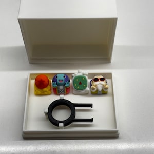 Könnte beinhalten: Weiße Box mit einem schwarzen Ring und vier kleinen Figuren. Die Figuren sind bunt und stellen Charaktere aus einer beliebten Anime-Serie dar.