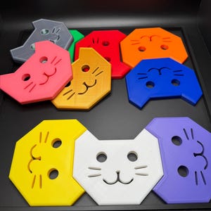 May include: A collection of colorful cat-shaped coasters in various colors, including pink, yellow, white, and blue. Each coaster features a simple cat face design with eyes, nose, and whiskers. The coasters are arranged on a black surface.