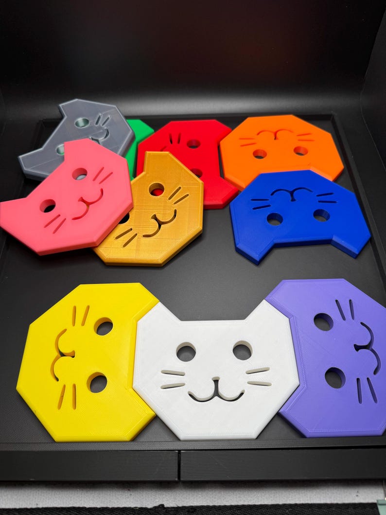 May include: A collection of colorful cat-shaped coasters in various colors, including pink, yellow, white, and blue. Each coaster features a simple cat face design with eyes, nose, and whiskers. The coasters are arranged on a black surface.