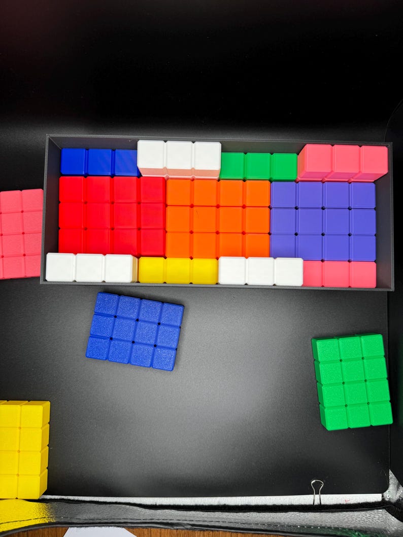 May include: A set of colorful square blocks in a black container. The blocks are arranged in a pattern and include red, orange, yellow, green, blue, purple, and white.