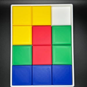 May include: A white tray with nine square compartments, each containing a different colored block. The blocks are yellow, red, green, and blue.