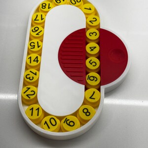 May include: A white plastic circular device with a red half-circle. The device has a series of yellow circles with black numbers 1 through 20 around the edge.