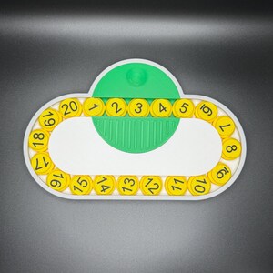 May include: A white plastic circular frame with yellow numbered circles around the edge. The numbers are 1 through 20. A green plastic piece with a small white knob is in the center of the frame.