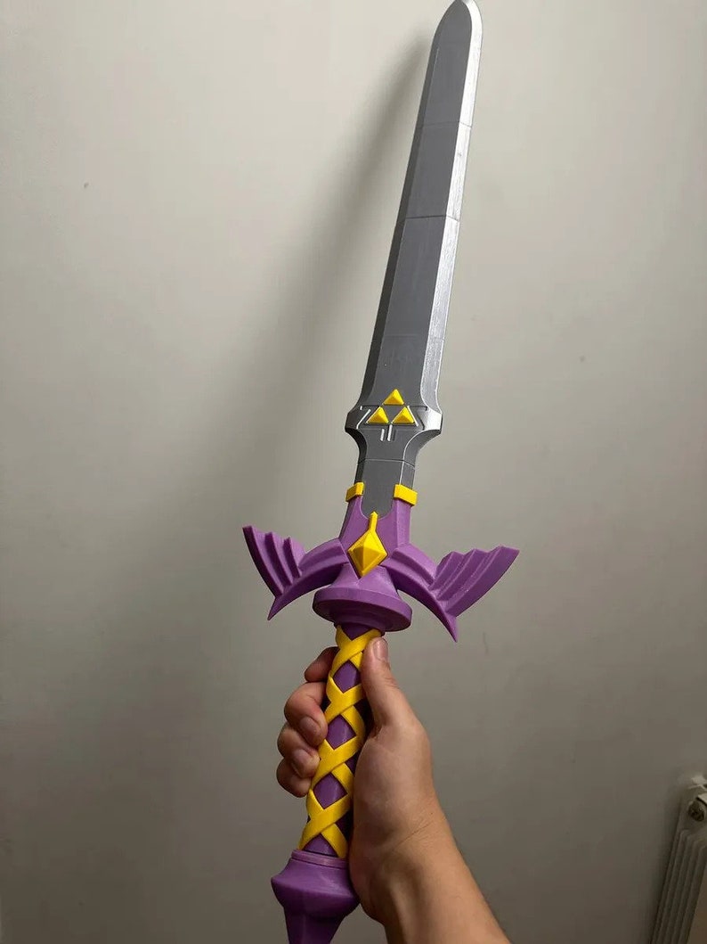 May include: A purple and yellow plastic sword with a silver blade and a triangular design. The sword has a detailed handle with a braided yellow design.