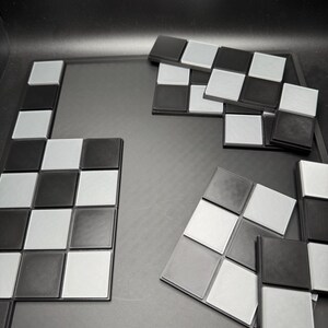 K&ouml;nnte beinhalten: Ein Schachbrettspiel in Schwarz und Silber mit einzelnen Schachfiguren. Das Spielbrett ist dunkelgrau und die Schachfiguren sind in einem Schachbrettmuster angeordnet. Die Figuren sind schwarze und silberne Quadrate.