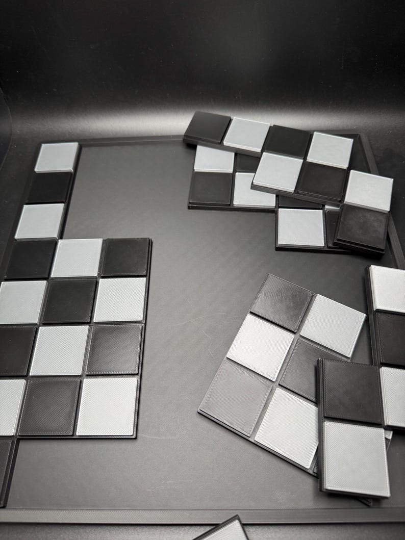 K&ouml;nnte beinhalten: Ein Schachbrettspiel in Schwarz und Silber mit einzelnen Schachfiguren. Das Spielbrett ist dunkelgrau und die Schachfiguren sind in einem Schachbrettmuster angeordnet. Die Figuren sind schwarze und silberne Quadrate.