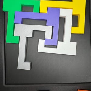 Op de afbeelding: Een puzzel met vijf stukken, elk in een andere kleur en vorm. De stukken zijn in een patroon op een zwart oppervlak gerangschikt. De kleuren zijn groen, geel, paars, wit en grijs.