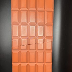 May include: A large, rectangular chocolate bar with a broken piece. The bar is divided into squares and has a deep orange colour.