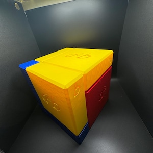 May include: A cube puzzle made of three different colored blocks. The top block is yellow, the front block is blue, and the side block is red. Each block has a number "3" printed on it.