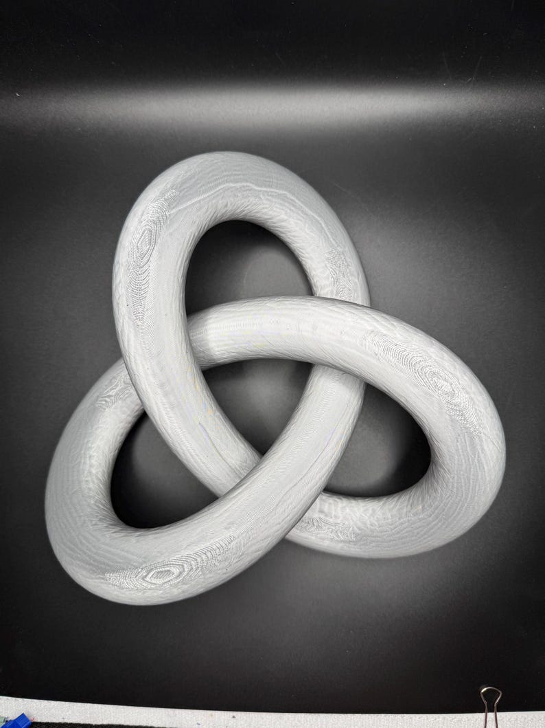 May include: A white, wooden, three-dimensional knot sculpture. The knot is made of three intertwined loops.