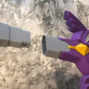 May include: A close-up of two plastic toy swords, one gray and one purple with a yellow accent. The swords are facing each other, with the tips of the blades almost touching.