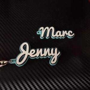 K&ouml;nnte beinhalten: Ein t&uuml;rkisfarbener Schl&uuml;sselanh&auml;nger mit den Namen "Jenny" und "Marc" in wei&szlig;er kursivschrift.