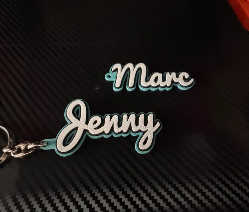 K&ouml;nnte beinhalten: Ein t&uuml;rkisfarbener Schl&uuml;sselanh&auml;nger mit den Namen "Jenny" und "Marc" in wei&szlig;er kursivschrift.