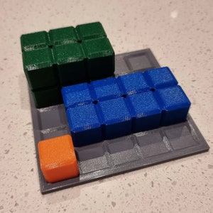 May include: A gray square game board with a green, blue, and orange cube on top. The cubes are arranged in a staggered pattern.