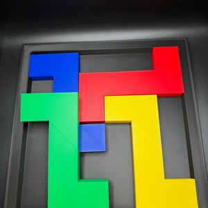 Op de afbeelding: Een kleurrijke puzzel met geometrische vormen in rood, blauw, groen en geel. De puzzelstukken zijn gerangschikt in een zwart frame, waardoor een visueel aantrekkelijk ontwerp ontstaat. De vormen grijpen in elkaar, wat wijst op een spel of educatief speelgoed.