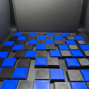 May include: A close-up of a board game with a grid of alternating black and blue squares. The squares are raised and have a textured surface. The game board is set against a dark background.