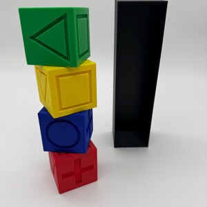 May include: Four colorful wooden blocks stacked on top of each other. The top block is green and has a triangle on it. The second block is yellow and has a square on it. The third block is blue and has a circle on it. The bottom block is red and has a plus sign on it. The blocks are next to a black rectangular box.