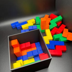 May include: A black box filled with colorful Tetris-like blocks. The blocks are red, orange, yellow, green, blue, and light blue. Some blocks are stacked on top of each other, while others are scattered around the box.