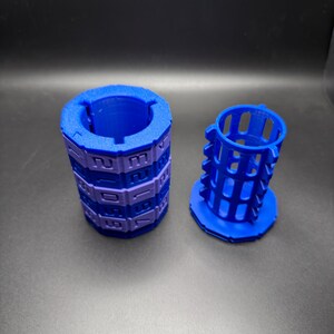 May include: Two blue and purple 3D-printed objects. One is a cylindrical puzzle with numbers, the other is a cage-like structure. The objects are on a dark surface.