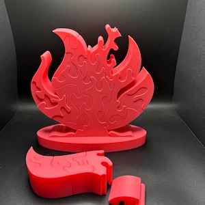 Puede incluir: Un rompecabezas de color rojo brillante con forma de llama que se mantiene en posici&oacute;n vertical sobre una base ovalada. El rompecabezas consta de varias piezas entrelazadas con bordes curvos y org&aacute;nicos. Dos piezas adicionales est&aacute;n separadas.