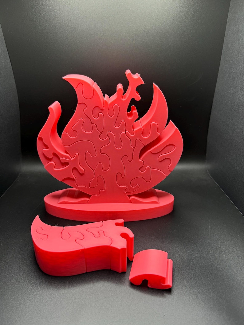Puede incluir: Un rompecabezas de color rojo brillante con forma de llama que se mantiene en posici&oacute;n vertical sobre una base ovalada. El rompecabezas consta de varias piezas entrelazadas con bordes curvos y org&aacute;nicos. Dos piezas adicionales est&aacute;n separadas.