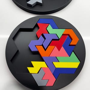 May include: A circular puzzle board with a hexagonal center. The puzzle board is black and features a variety of colorful arrow-shaped pieces. The pieces are red, orange, yellow, green, blue, purple, and gray.
