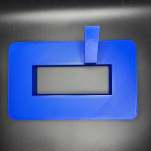 May include: A blue plastic frame with a rectangular cutout. The frame has a small blue plastic piece attached to the top.