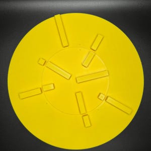 May include: Bright yellow circular object with rectangular cutouts arranged around a central circle. The object is set against a black background, creating a high-contrast image. The cutouts are evenly spaced.