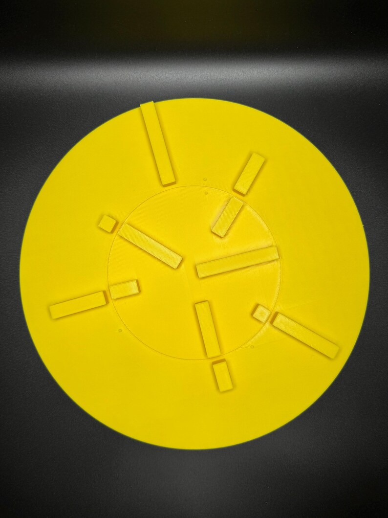 May include: Bright yellow circular object with rectangular cutouts arranged around a central circle. The object is set against a black background, creating a high-contrast image. The cutouts are evenly spaced.
