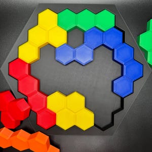 May include: A hexagonal puzzle with a black frame and colorful pieces. The pieces are arranged in a curved shape, with yellow, red, blue, and green pieces visible. The puzzle is made of plastic and is designed to be challenging and fun.