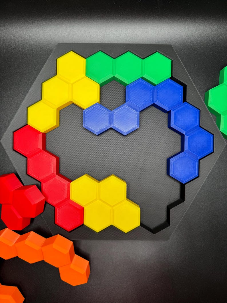 May include: A hexagonal puzzle with a black frame and colorful pieces. The pieces are arranged in a curved shape, with yellow, red, blue, and green pieces visible. The puzzle is made of plastic and is designed to be challenging and fun.