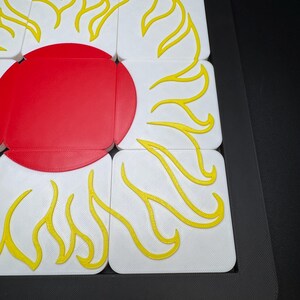 May include: A close-up of a puzzle featuring a red sun with yellow flames on white squares. The puzzle is set in a black frame. The sun has a face with a smiling mouth and closed eyes.