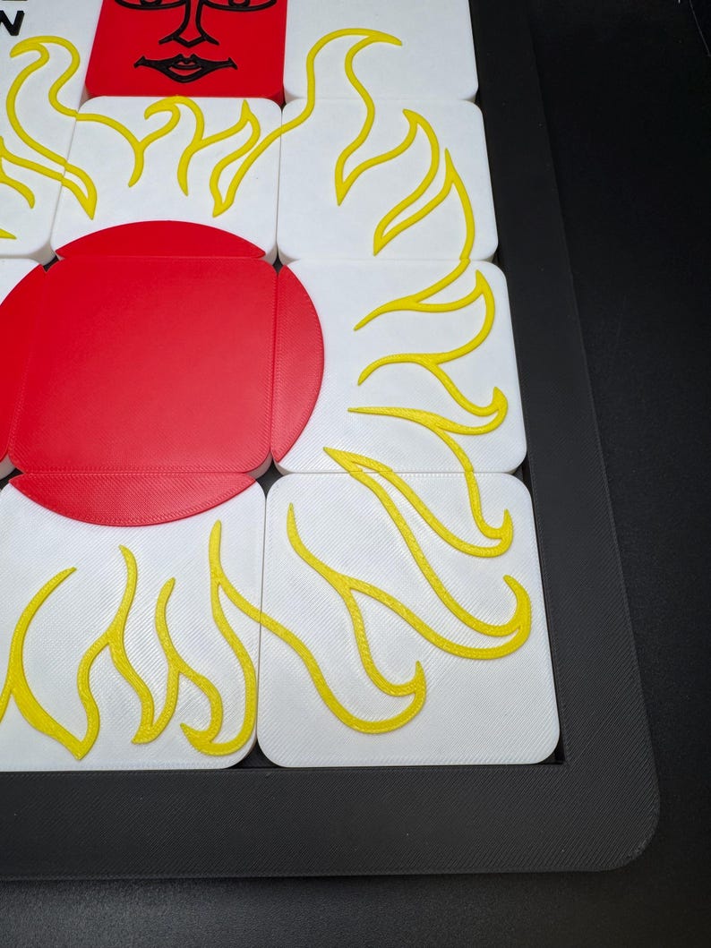 May include: A close-up of a puzzle featuring a red sun with yellow flames on white squares. The puzzle is set in a black frame. The sun has a face with a smiling mouth and closed eyes.