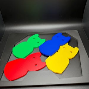 May include: Four colorful cat-shaped cutouts in red, green, blue, and yellow. Each cutout has two small circular holes and a notched top, resembling cat ears. The cutouts are arranged on a dark gray surface.