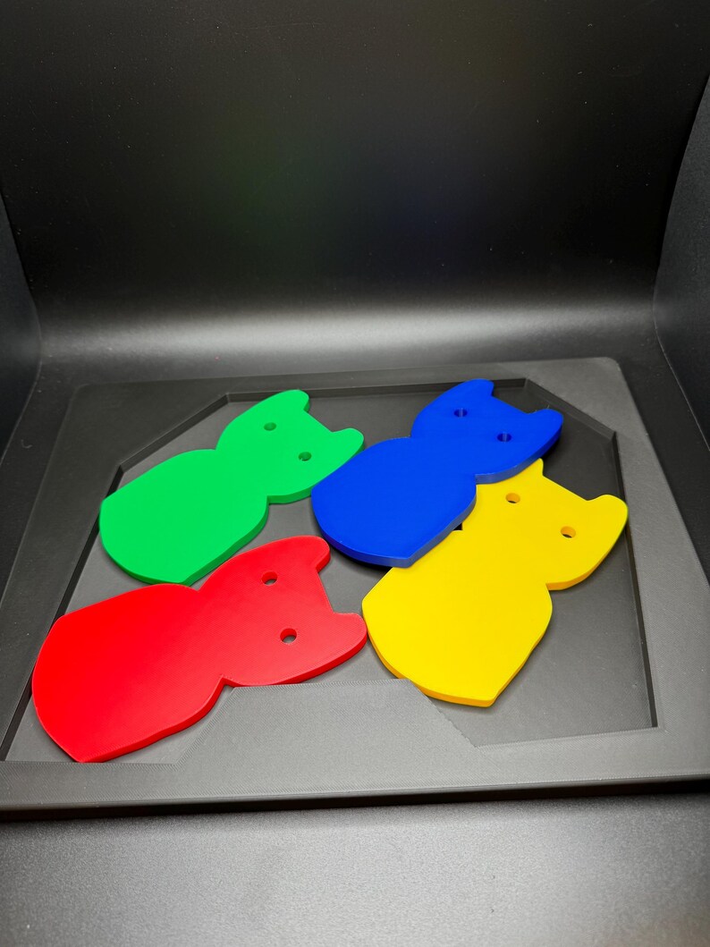 May include: Four colorful cat-shaped cutouts in red, green, blue, and yellow. Each cutout has two small circular holes and a notched top, resembling cat ears. The cutouts are arranged on a dark gray surface.