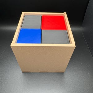 May include: A wooden box with four compartments, each containing a colored block. The blocks are red, blue, and two gray blocks.