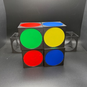 May include: Four black cubes arranged together, each with a coloured circle on one side. The circles are red, green, blue, and yellow. The cubes are arranged in a 2x2 pattern. The background is black.