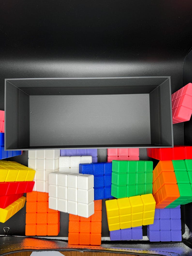 May include: A black box filled with colorful building blocks. The blocks are made of plastic and have a grid pattern. The blocks are in various colors, including red, orange, yellow, green, blue, purple, and white.