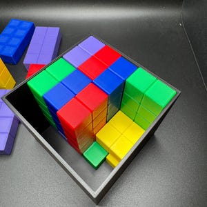 May include: A colorful puzzle game featuring a cube-shaped frame filled with various colored blocks. The blocks are arranged in a grid pattern, with colors including red, green, blue, yellow, and purple. Additional blocks are visible outside the frame.