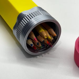 May include: A yellow pencil-shaped container with a silver lid, open to reveal a collection of colored pencils inside.