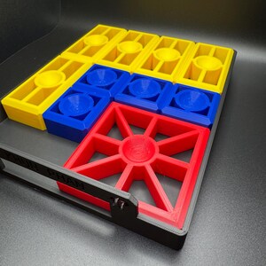 Puede incluir: Un tablero de juego impreso en 3D con piezas amarillas, azules y rojas. El tablero tiene un marco negro con las palabras "FUN PLAN" impresas en &eacute;l.