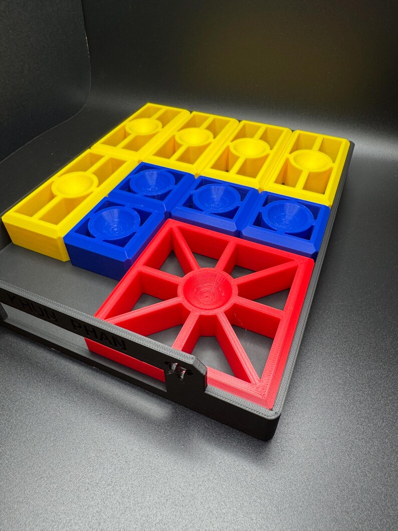 Puede incluir: Un tablero de juego impreso en 3D con piezas amarillas, azules y rojas. El tablero tiene un marco negro con las palabras "FUN PLAN" impresas en &eacute;l.