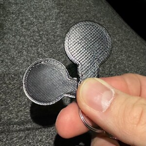 May include: A silver-coloured keychain featuring two circular elements connected by a stem. The surface has a textured pattern. The keychain is attached to a silver ring. The item is likely made of metal.