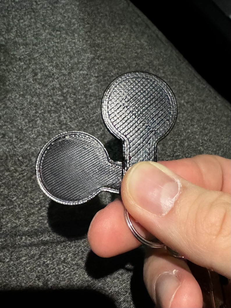 May include: A silver-coloured keychain featuring two circular elements connected by a stem. The surface has a textured pattern. The keychain is attached to a silver ring. The item is likely made of metal.