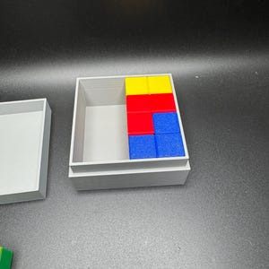 Puede incluir: Una caja gris con la tapa abierta, que revela un conjunto de bloques de madera de colores. Los bloques est&aacute;n dispuestos en un patr&oacute;n, con bloques amarillos, rojos y azules visibles. Los bloques est&aacute;n dispuestos en un patr&oacute;n, con bloques amarillos, rojos y azules visibles.