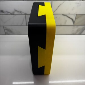 May include: A black and yellow box with a lightning bolt design. The box is made of a hard plastic material and has a smooth finish.